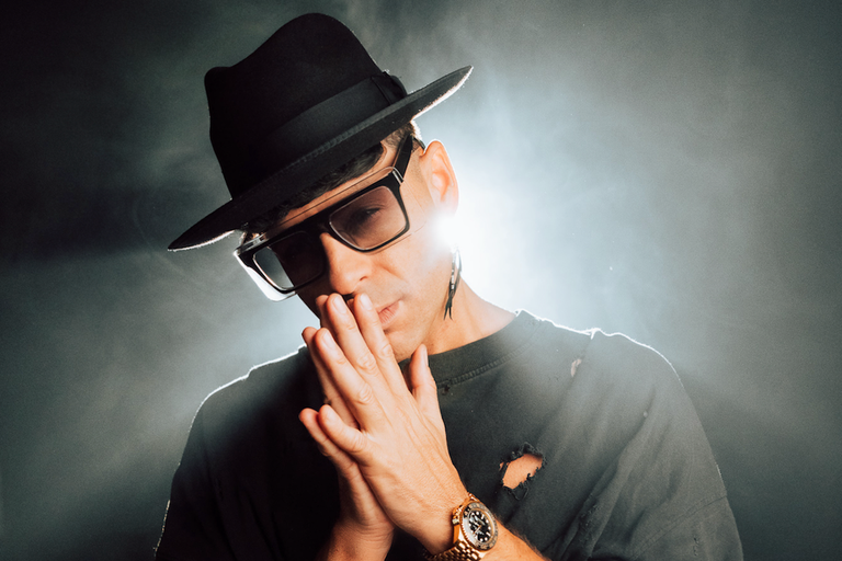 Australian DJ Timmy Trumpet wearing a black fedora and sunglasses, posing with hands clasped in front of a hazy backdrop.
